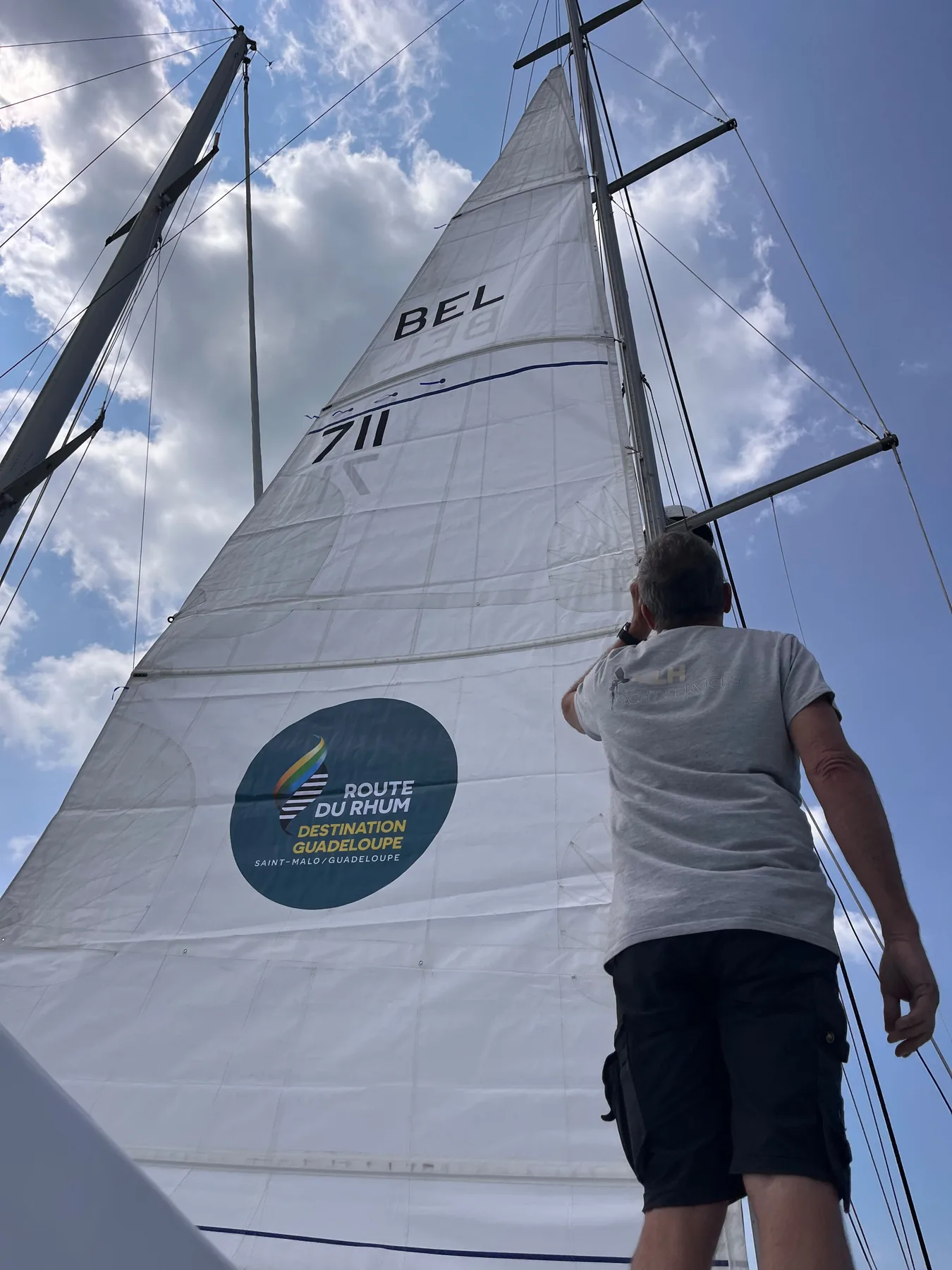 Amaury regarde vers la grand-voile portant le numéro BEL 711 et l’autocollant de la Route du Rhum
