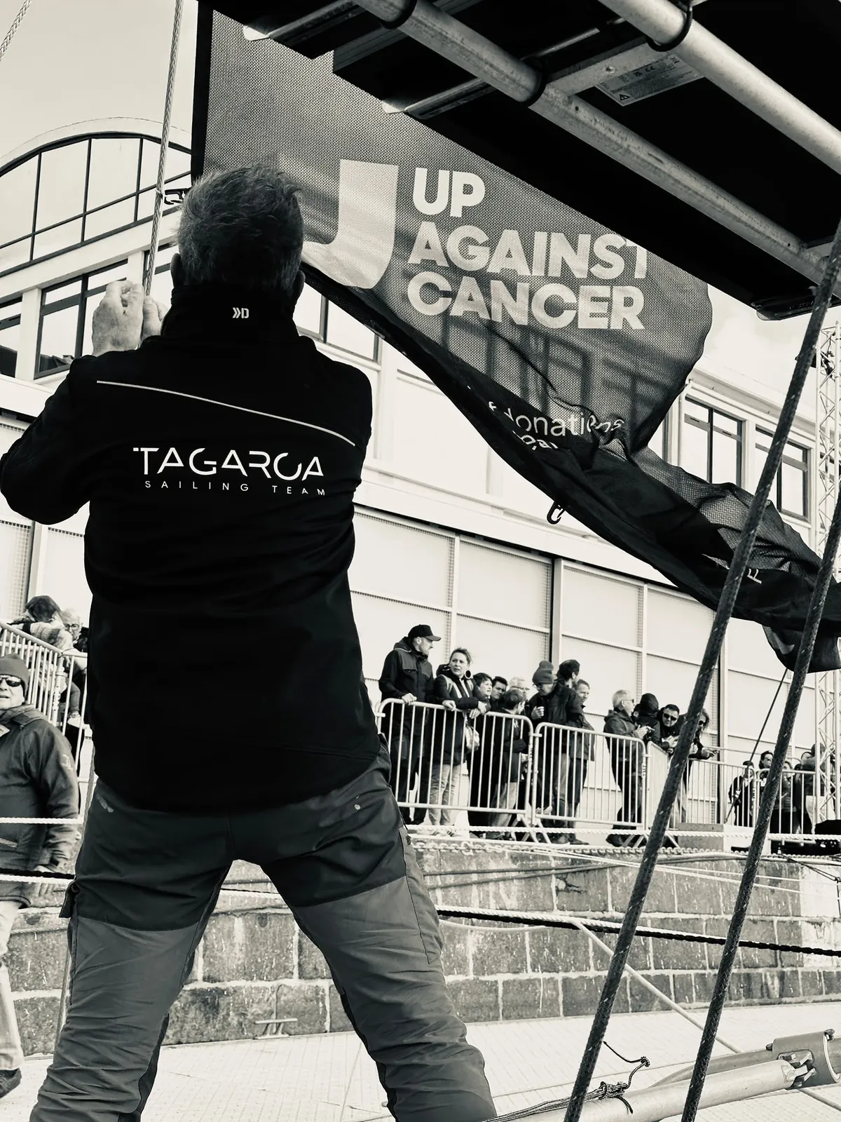 Amaury de dos dans sa veste Tagaroa, face à la bannière d’Up Against Cancer et au public sur le quai